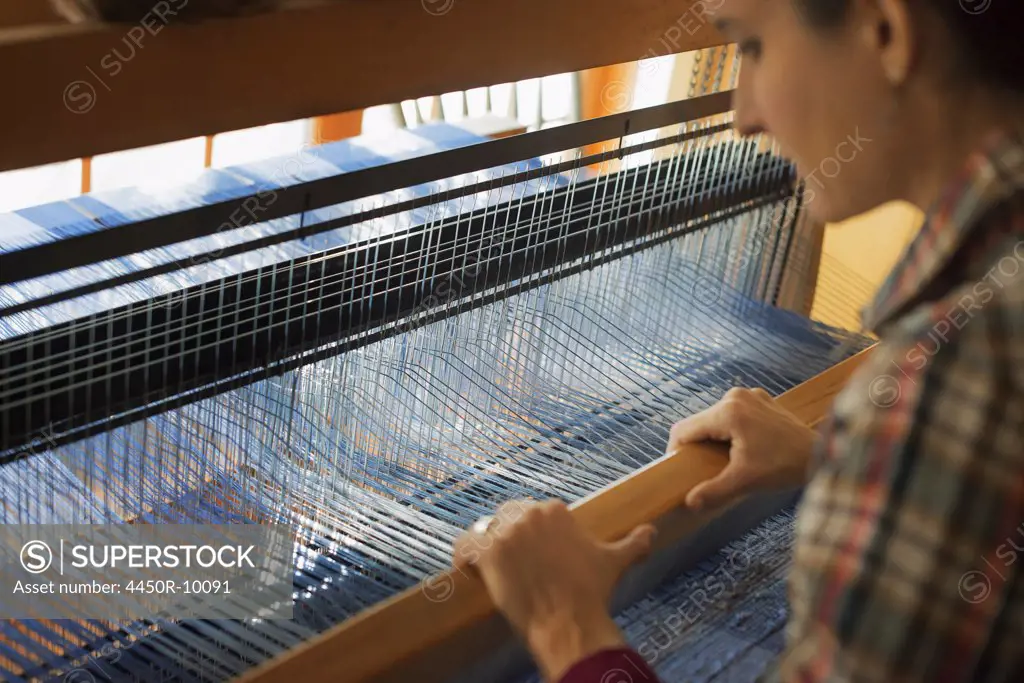 A woman seated at a wooden handloom creating a handwoven woollen fabric, with a blue and white pattern. Woodstock, New York, USA