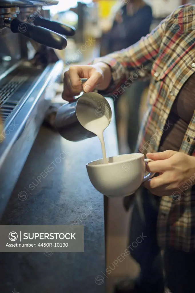 A person, barista, making coffee, and pouring frothed milk from a jug into a cup for a cappuccino. Coffee shop.Kingston, New York, USA
