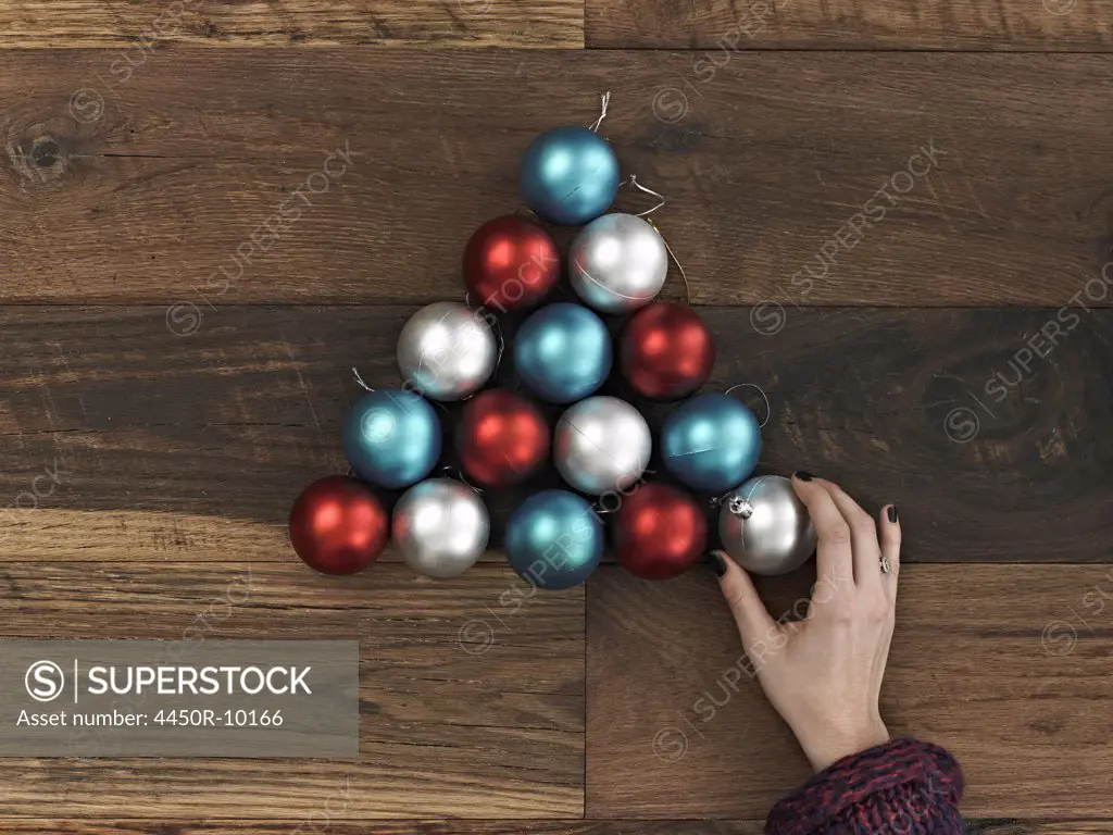 A collection of blue, red and silver ornaments arranged in a triangular shape on a wooden board. A Christmas tree shape. A person's hand placing the final ball.New York city, USA