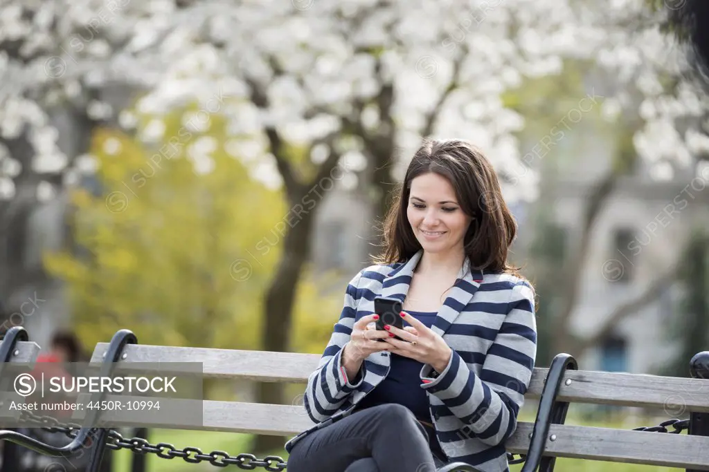 Outdoors in the city in spring time. New York City park. White blossom on the trees. A woman sitting on a bench holding her mobile phone. New York city, USA