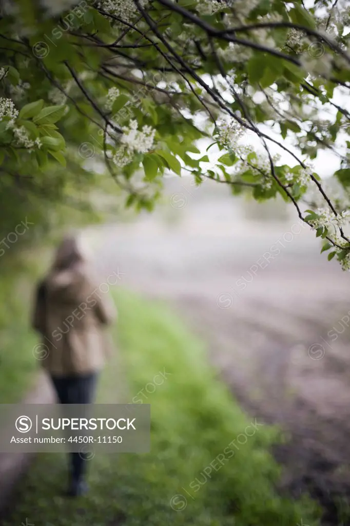 A woman in a brown hooded coat and hat walking along a path beside a ploughed field. Rome, Italy