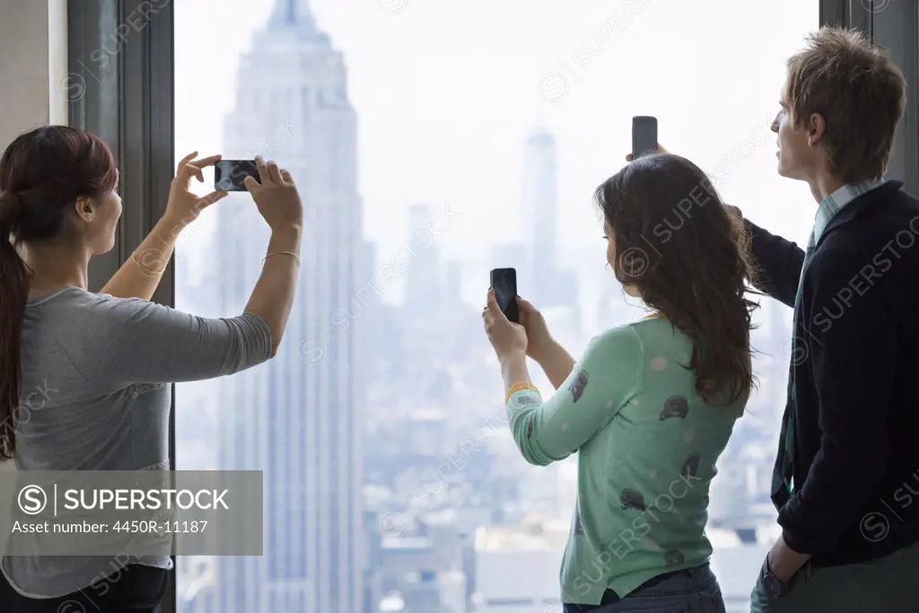 Urban lifestyle. Three people standing on an observation deck, using their phones to take images of the view over the city.  New York city, USA