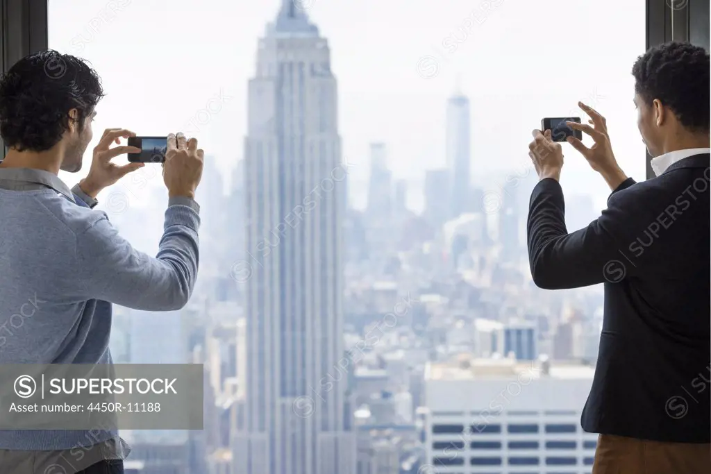 Urban lifestyle. Two young men using their phones to take images of the city from an observation platform overlooking the Empire State Building.  New York city, USA