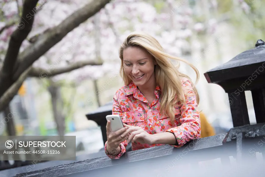 City life in spring. A young woman with long blonde hair sitting in a city park, looking at her smart phone.  New York city, USA