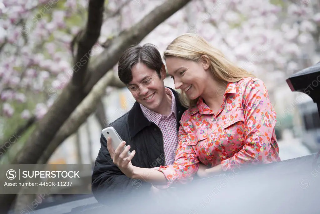 City life in spring. Young people outdoors in a city park. A young woman and man sitting side by side looking down at a smart phone.  New York city, USA