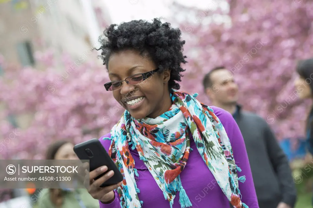 A group of people under the cherry blossom trees in the park. A young woman smiling and checking her phone. New York city, USA