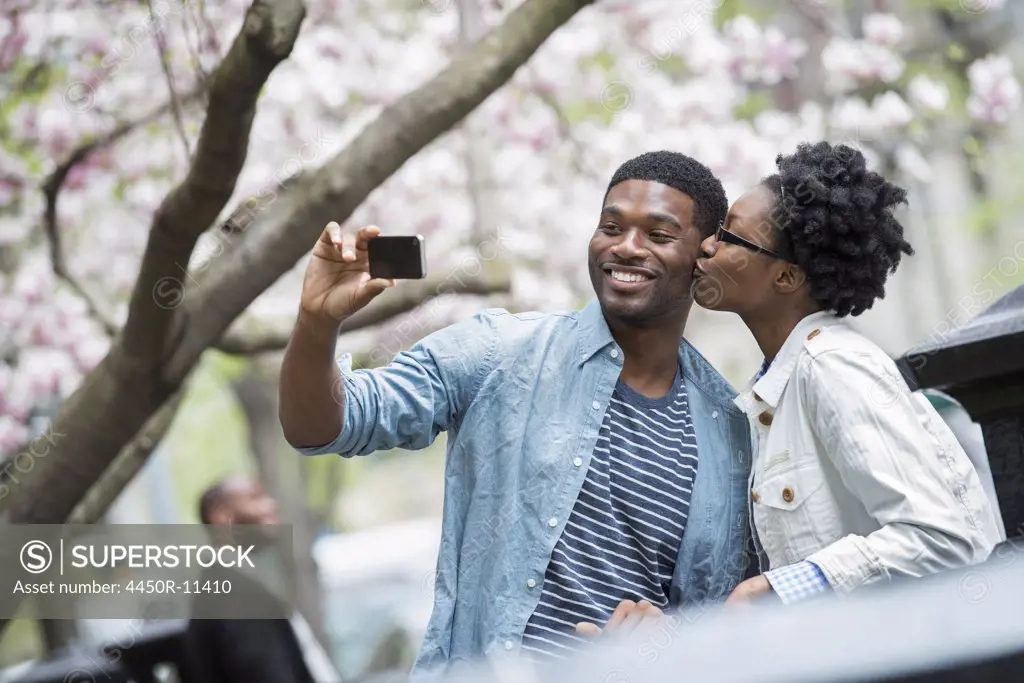 Outdoors in the city in spring. An urban lifestyle. A woman kissing a man and taking a photograph with a handheld mobile phone. New York City, USA