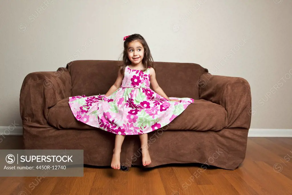 A 3 year old girl with long brown hair in a pink flowered cotton dress with the skirt spread out, sitting on a brown sofa.  Sante Fe, New Mexico, USA