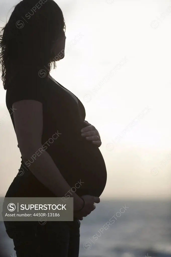 Silhouette of a heavily pregnant woman standing on a beach, with one hand resting on the top of her belly and one supporting it. England. 10/18/2011