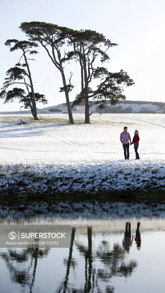 A couple standing in a snow-covered field by a river. Trees in background. England. 12/20/2008