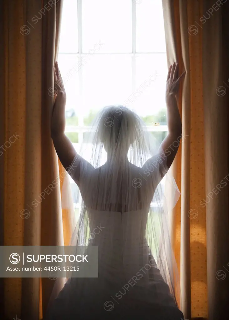 A woman in a wedding dress, wearing a light veil standing at a window with her arms up. England. 05/26/2012