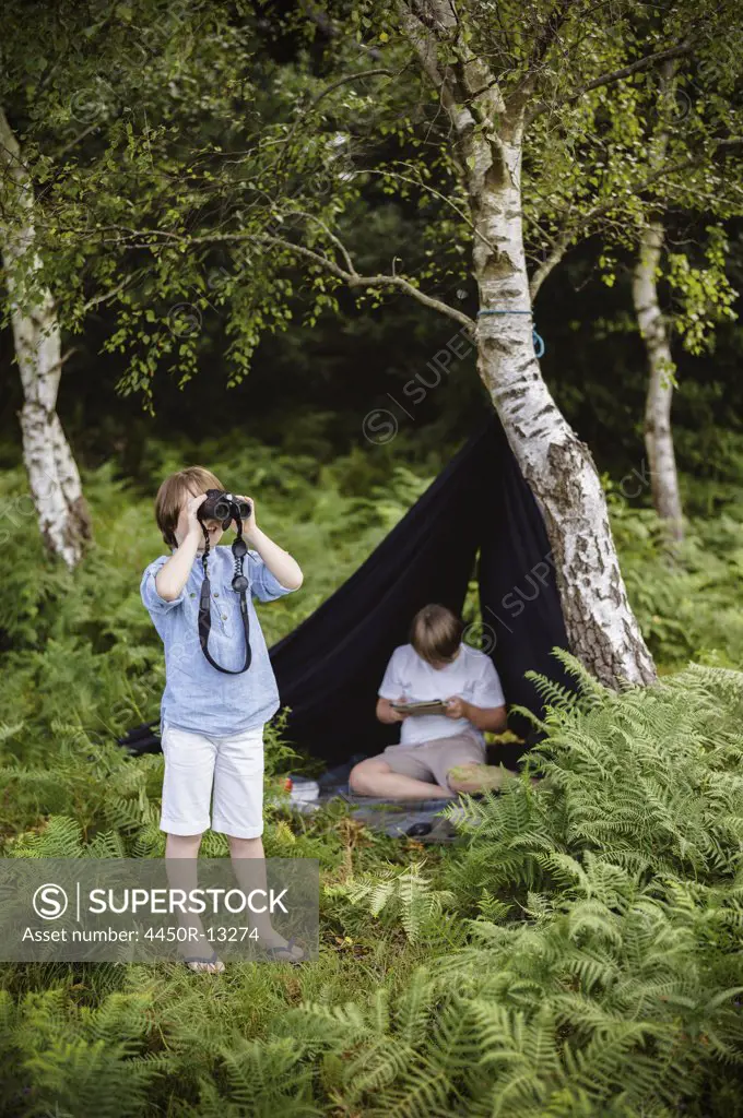 Two boys camping in New Forest. One sitting under a black canvas shelter.  One boy looking through binoculars. Hampshire, England. 08/06/2013