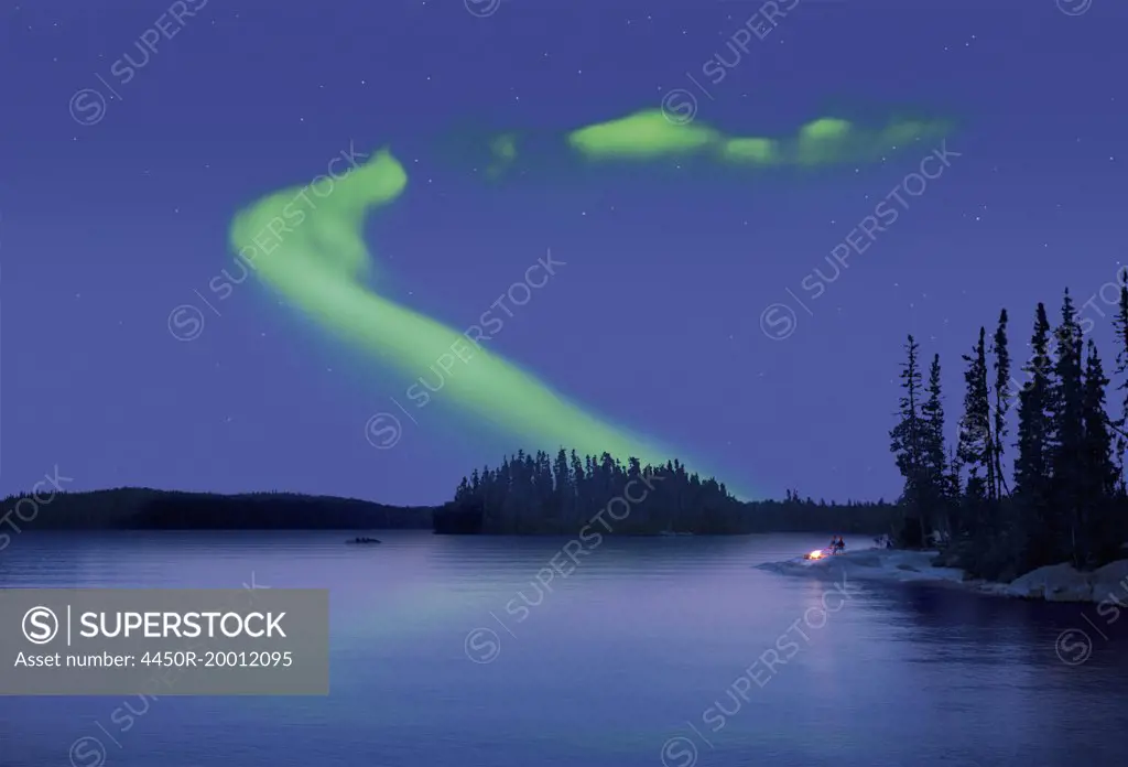 The Northern Lights in the night sky, the Aurora borealis above a calm lake. Two people by a fire on the lake shore.
