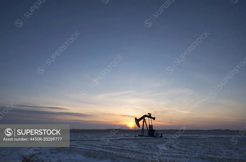 An oil drilling rig and pumpjack on a flat plain in the Canadian oil fields at sunset.