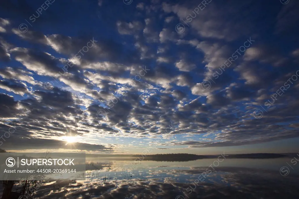 A dramatic cloud pattern across the sky and the sun setting over the waters of a lake.