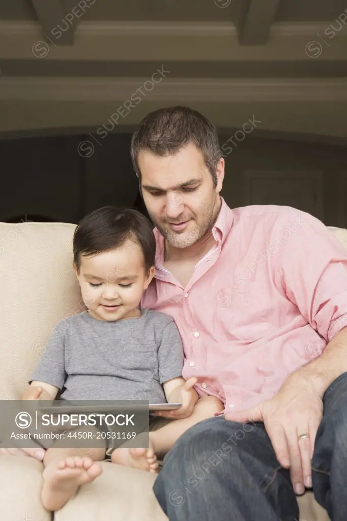 Smiling man sitting on a sofa with his young son, looking at a digital tablet.