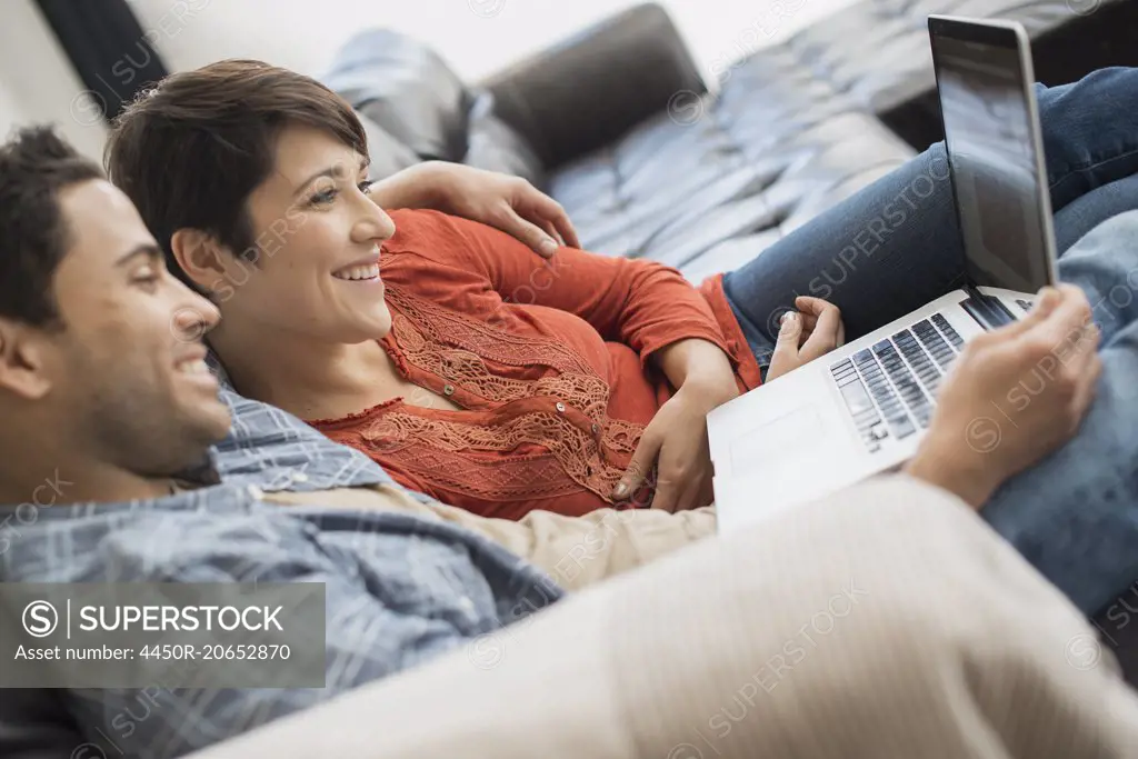A man and woman sitting on a sofa, looking at the screen of a laptop.