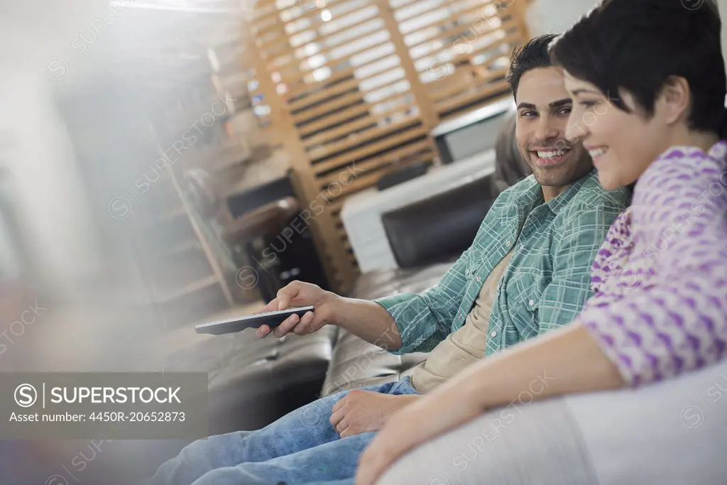 A couple, man and woman sitting on a sofa at home, one holding a remote control device.