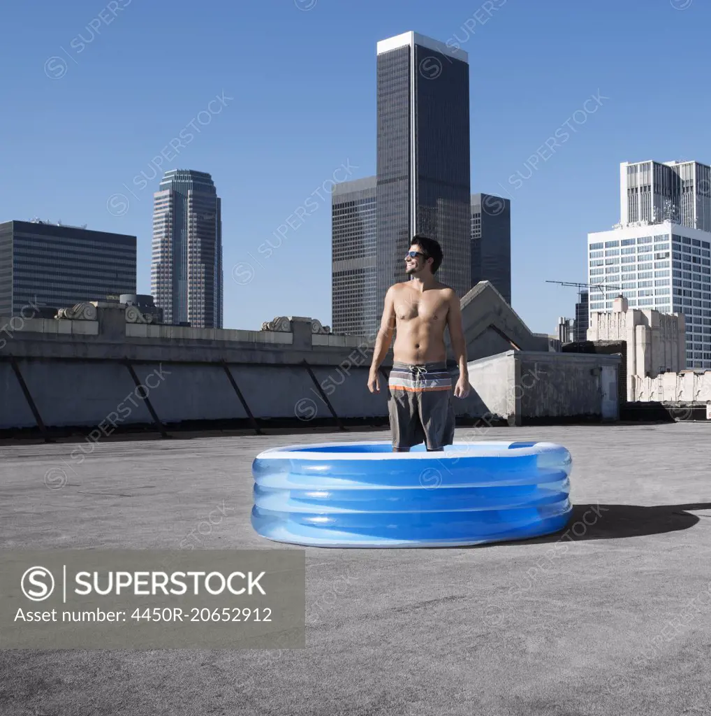 A man standing in a small inflatable water pool on a city rooftop.