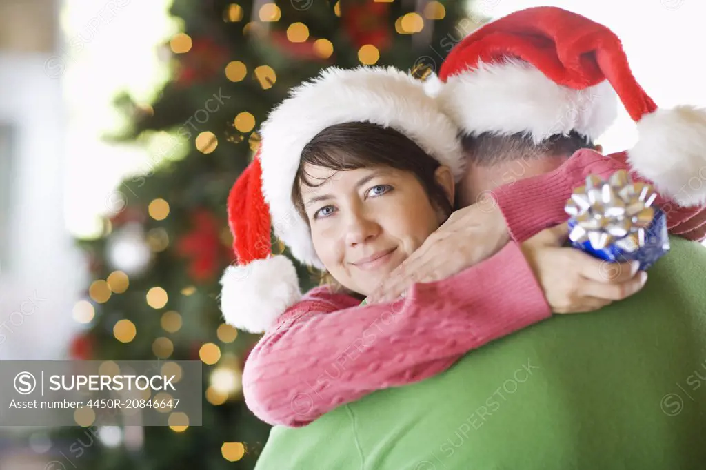 A couple wearing Santa hats hugging in front of a Christmas tree.