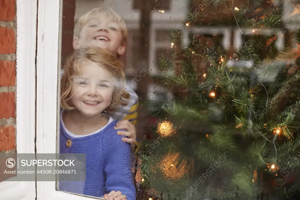 Two children, a boy and girl, looking out of a window at home beside a Christmas tree.