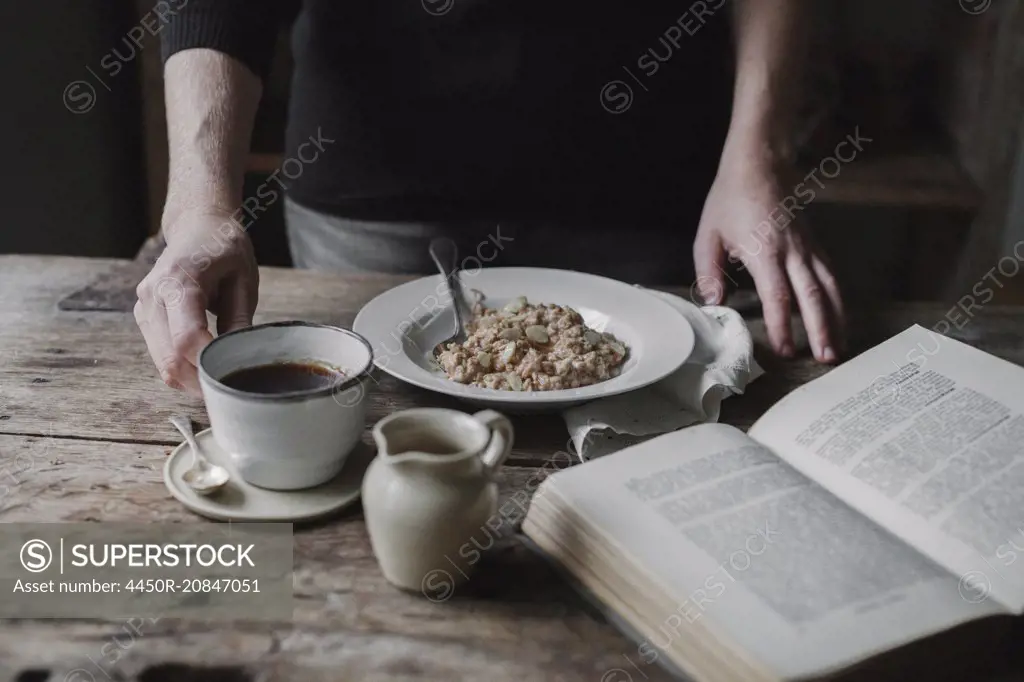 A person at a table with a cup of coffee, bowl of muesli and an open book.