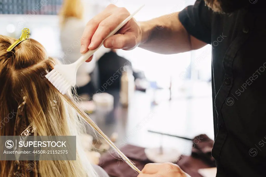 A hair colourist, a man using a paintbrush to cover sections of a woman's blonde hair.