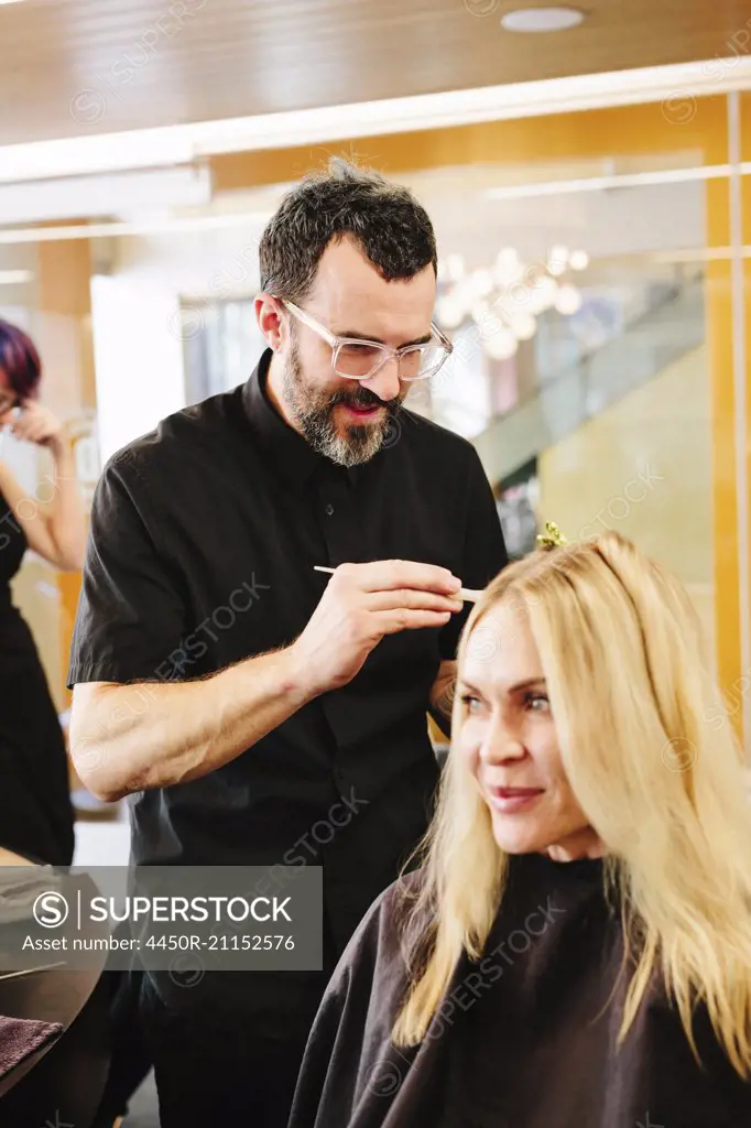 A hair colourist, a man using a paintbrush to cover sections of a woman's blonde hair.