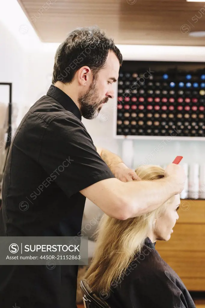 A mature man, a hair stylist, colourist, working on a woman's hair, applying hair colour to the centre parting.