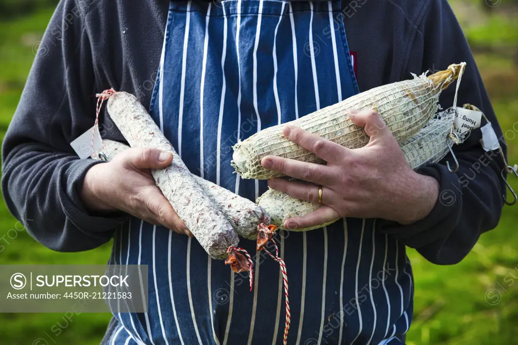 Close up of a butcher wearing a striped blue apron, standing outdoors, holding a selection of salamis.
