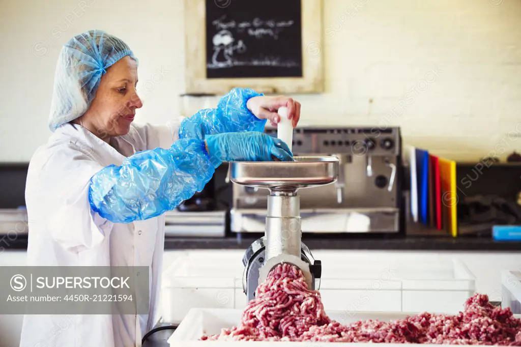 Woman working in a butchery, wearing protective clothes and gloves, putting minced meat into a meat grinder.