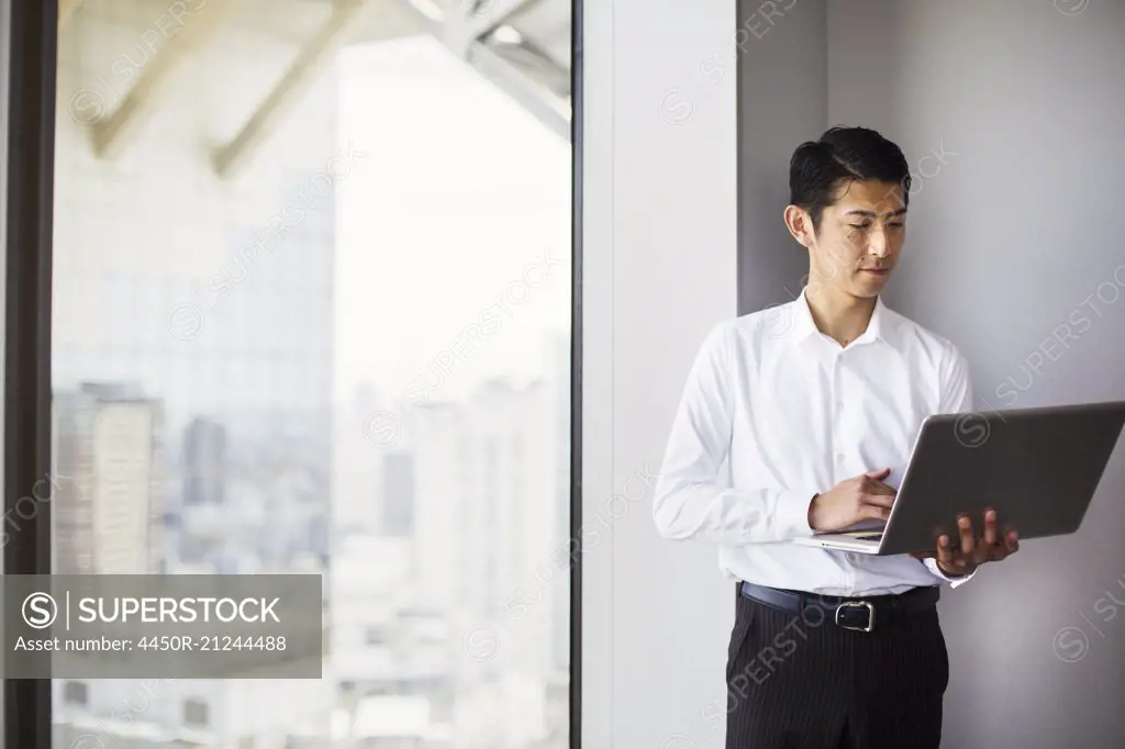A business man standing by a window with a city view, using his laptop.