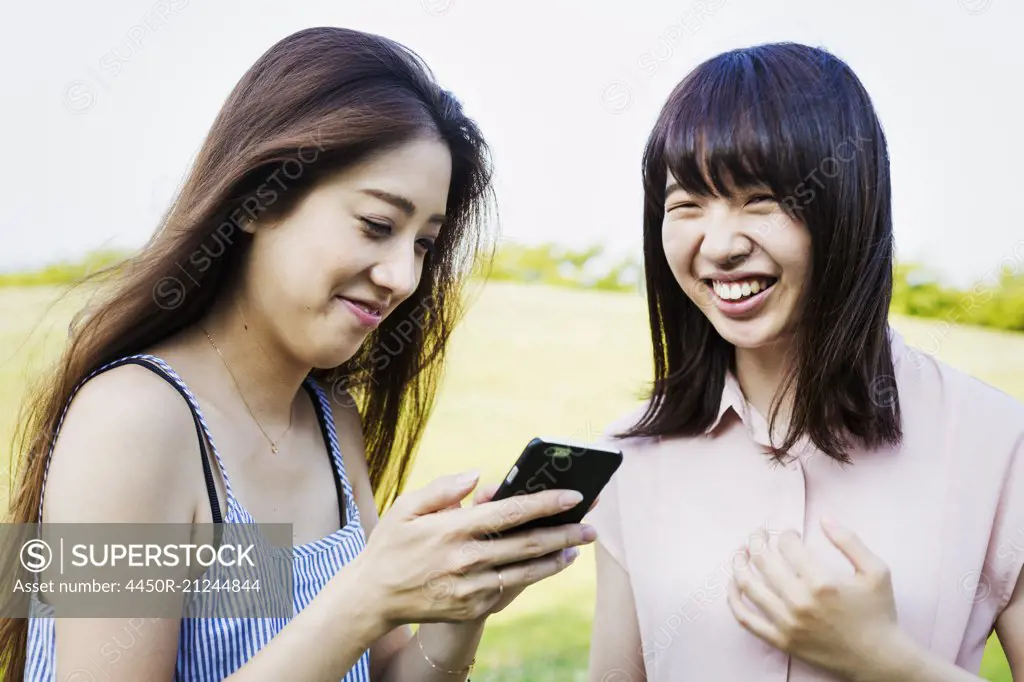 Two smiling young women with long brown hair, holding a mobile phone.
