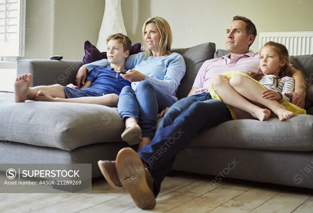 A family of four people, parents and a girl and boy, seated on the sofa together, watching television.