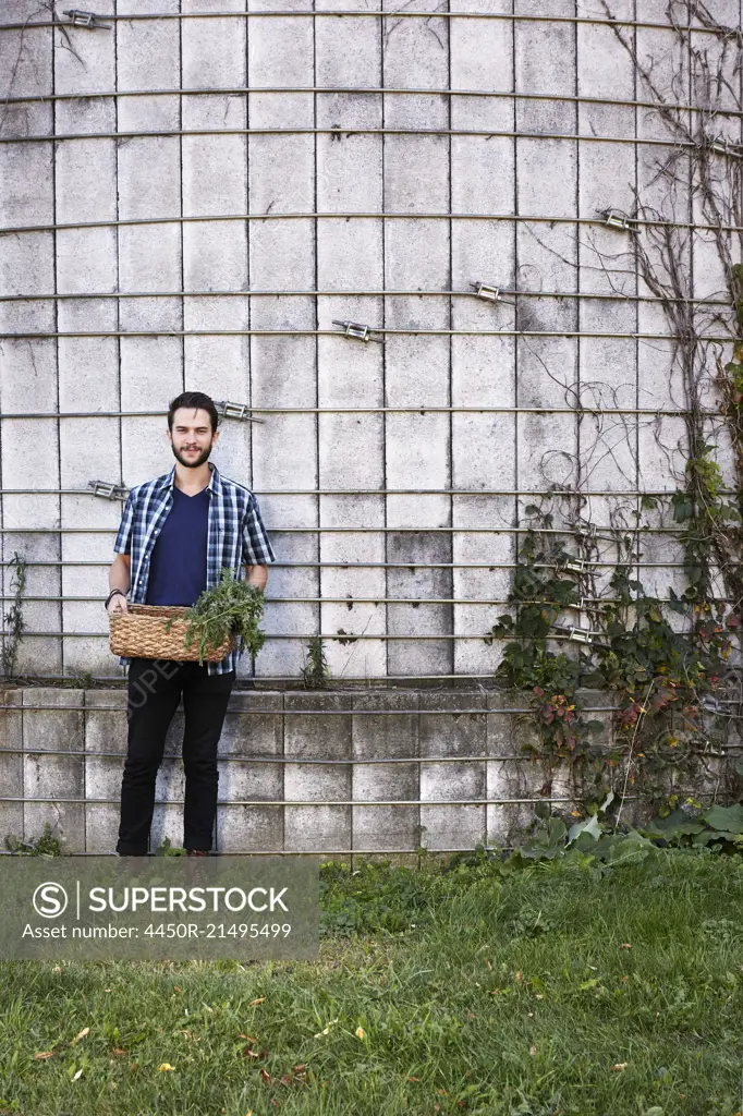 A young man in working clothes standing in front of a wall with plants growing up a trellis, holding a basket of crops.