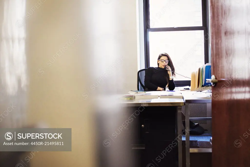 A young woman sitting at a desk in an office holding a telephone to her head.