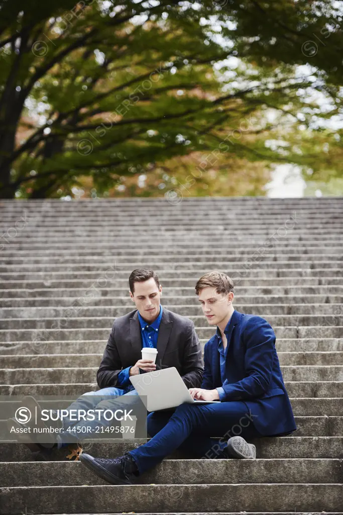 Two young men sitting on a flight of steps outdoors, looking at a laptop together.