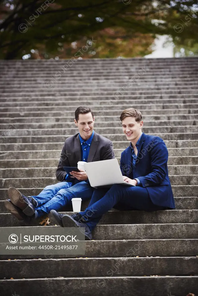 Two young men sitting on a flight of steps outdoors looking at a laptop together.