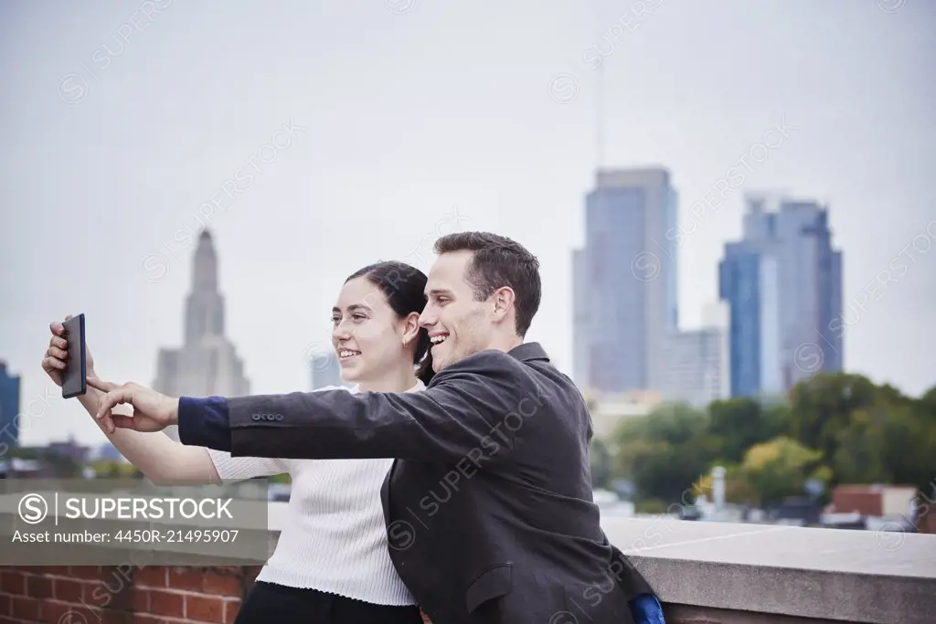 A young woman and a young man standing on a rooftop looking at a cellphone together.