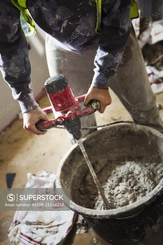 A builder mixing plaster using an electric mixer in a bucket on a construction site.
