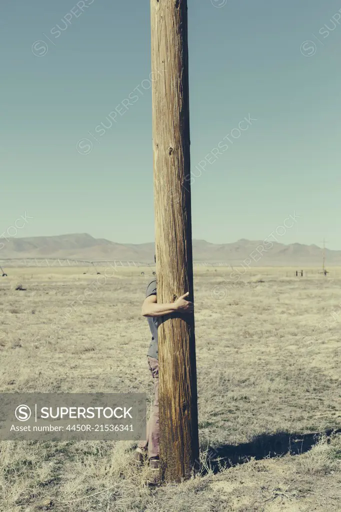Man with his arms around a wooden utliities pole, clinging to or hugging the post in a flat open landscape.