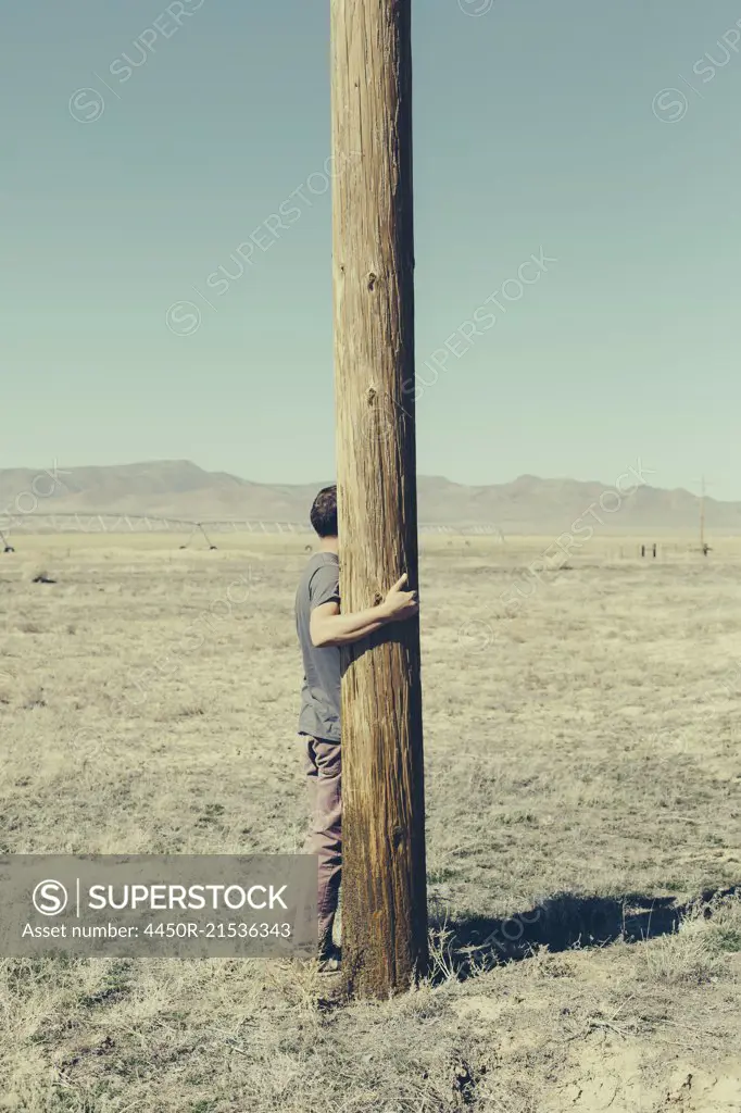 Man with his arms around a wooden utliities pole, clinging to or hugging the post in a flat open landscape.