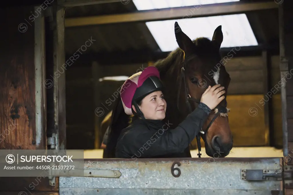 Woman wearing a riding helmet and brown horse standing in a box stall in a stable.