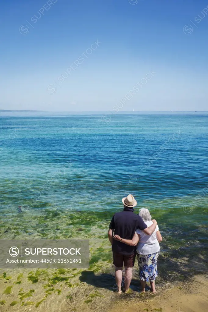 Rear view of elderly couple standing with arms around shoulders looking out to sea across the vivid clear blue water.