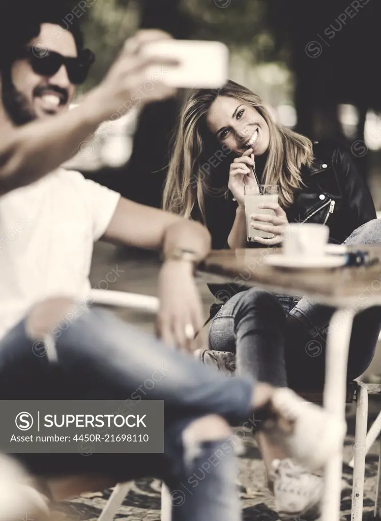 Bearded man wearing sunglasses and woman with long blond hair sitting outdoors at a table in a cafe, man holding smartphone, smiling and taking selfie.
