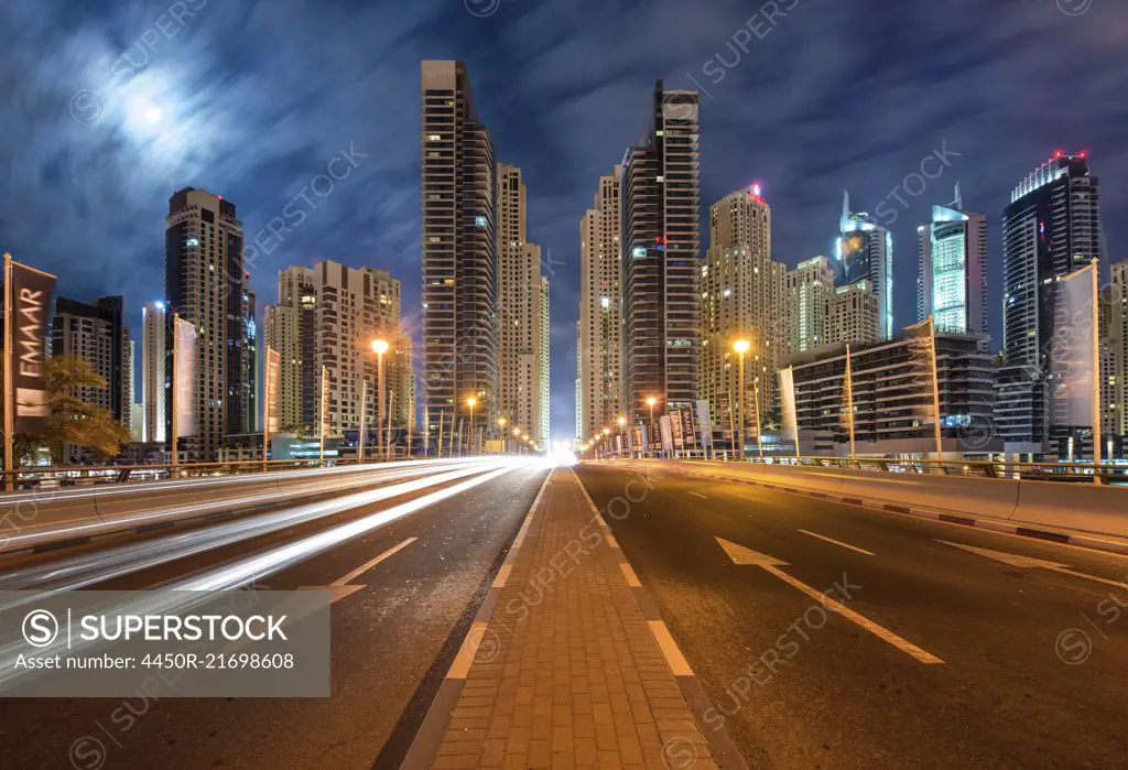 Cityscape with illuminated skyscrapers in Dubai, United Arab Emirates at dusk, highway in foreground.