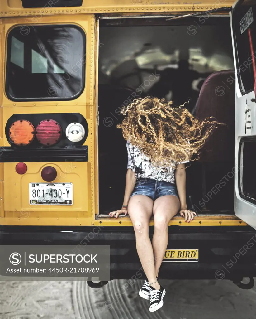 A young woman with long curly hair over her face sitting on the tailgate of a school bus.