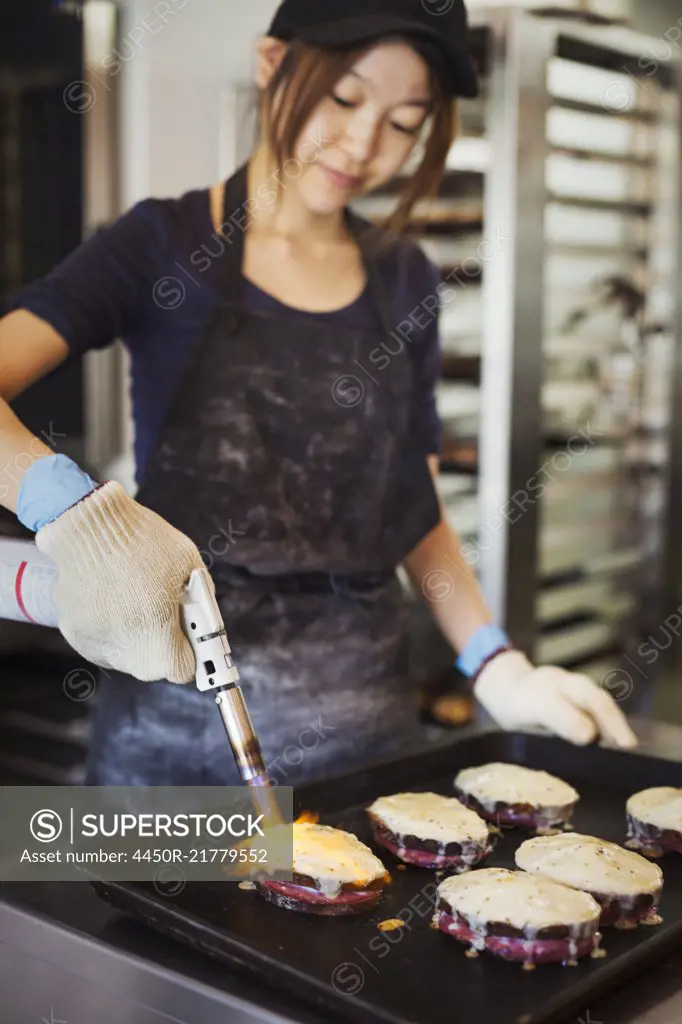Woman working in a bakery, wearing oven gloves, using blowtorch, melting cheese on sandwiches.