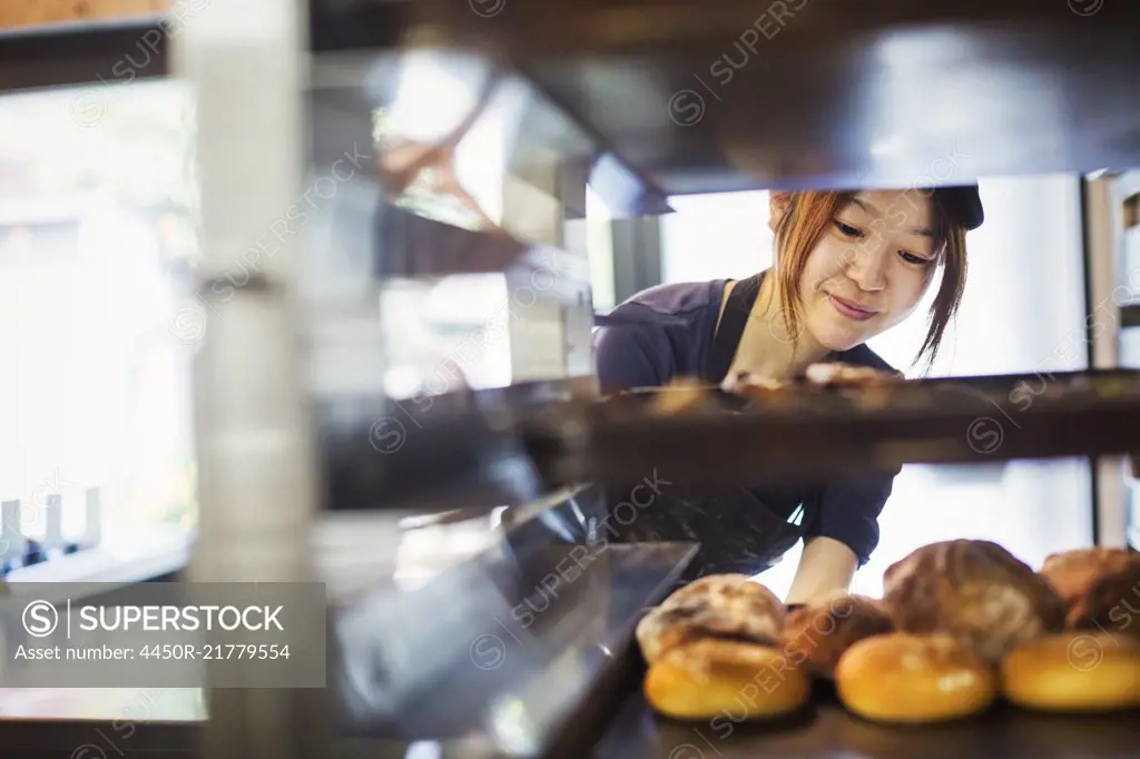 Woman working in a bakery, placing large trays with freshly baked rolls on a trolley.