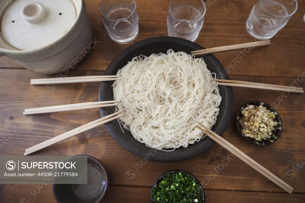 High angle close up of bowl with noodles and pairs of wooden chopsticks on a table.